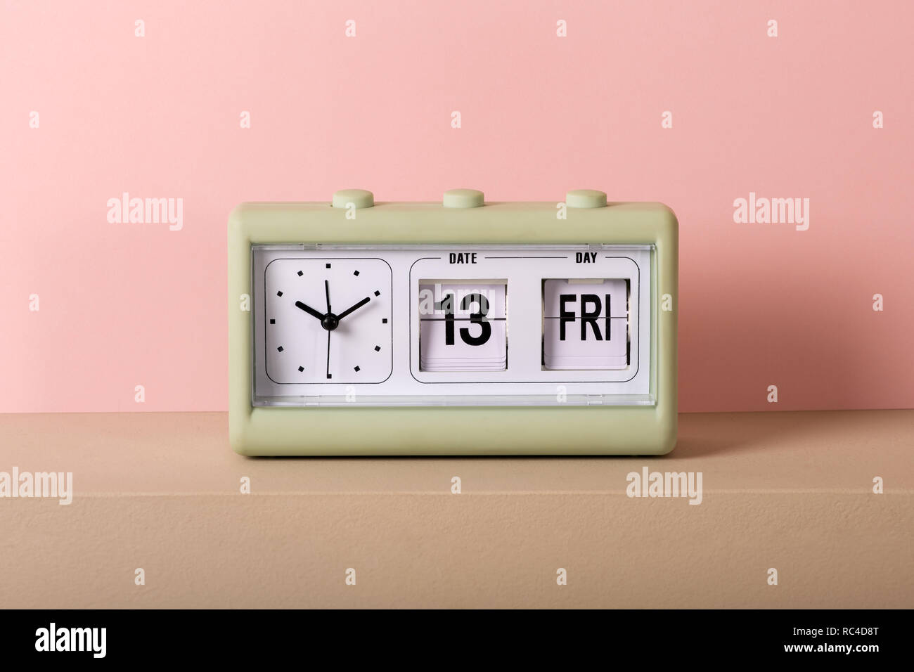 Small green vintage clock with white face and calendar showing Friday 13th. Viewed from the front in close-up, against pale pink background Stock Photo