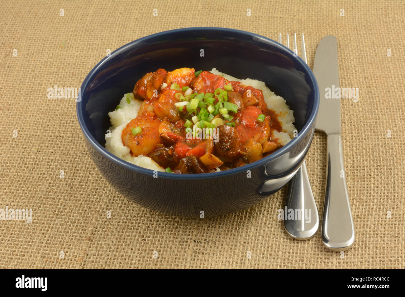 Shrimp and grits with mushrooms and red peppers in blue bowl with knife and fork on burlap Stock Photo