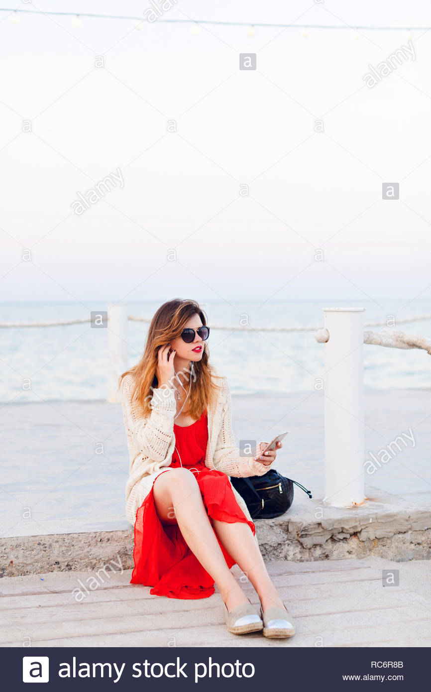 Beautiful Girl In Red Dress And White Jacket Sits On A Pier