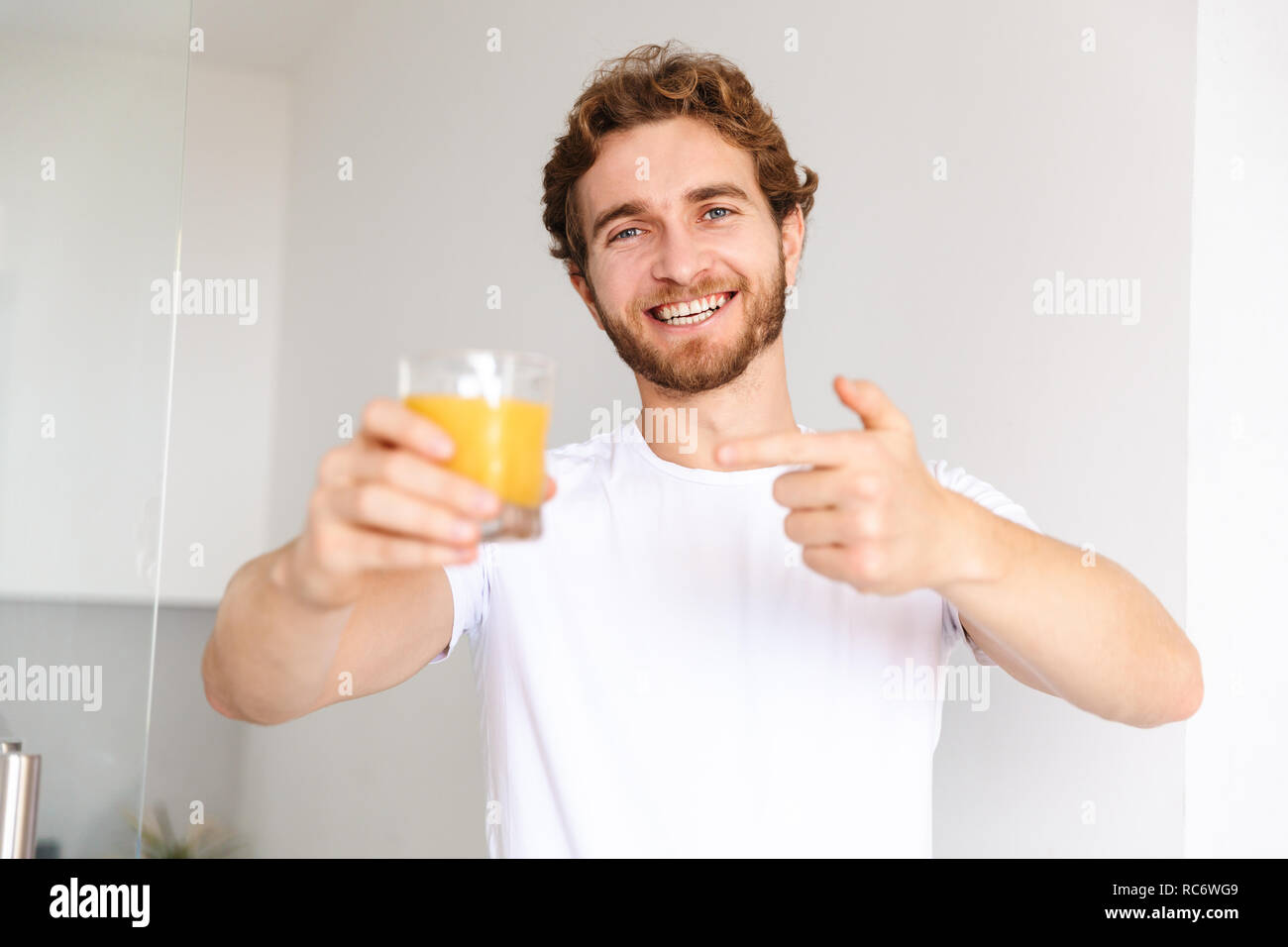 Image of a handsome young bearded man at home drinking juice showing thumbs up. Stock Photo