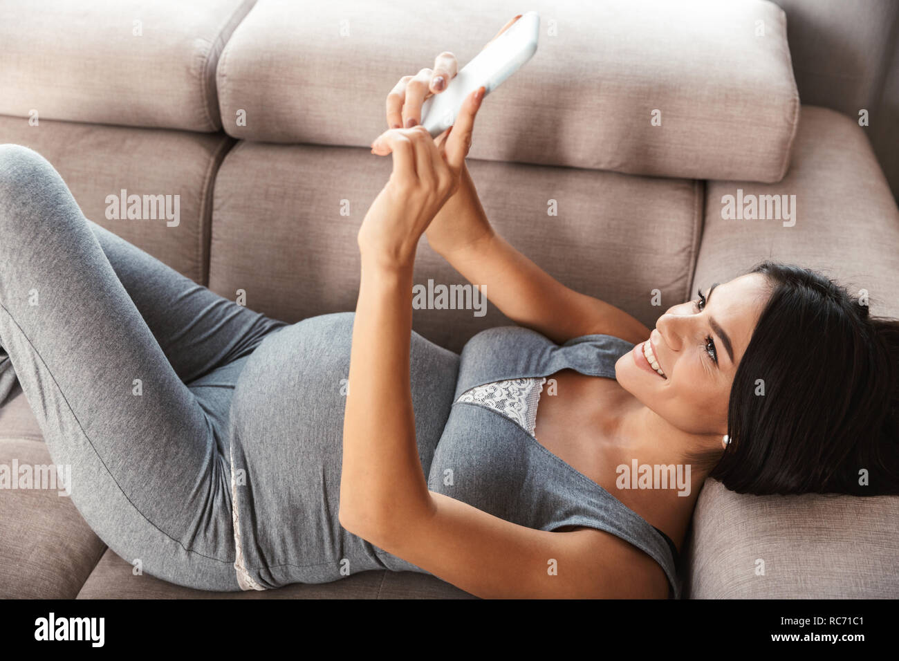Happy young pregnant woman laying on a couch at home, taking a selfie Stock Photo