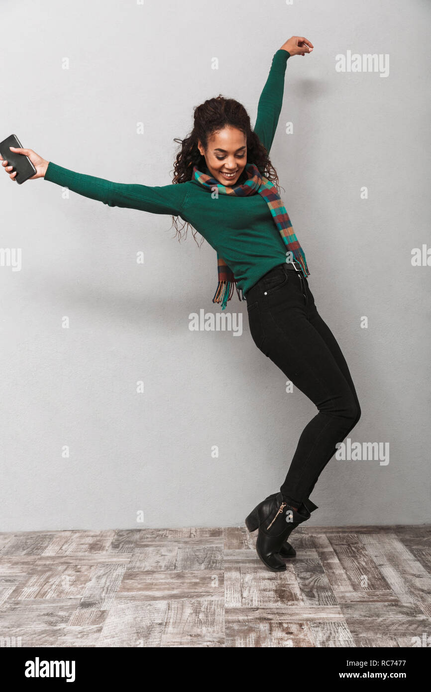 Full length portarit of a pretty african woman wearing scarf jumping over gray background, holding mobile phone Stock Photo