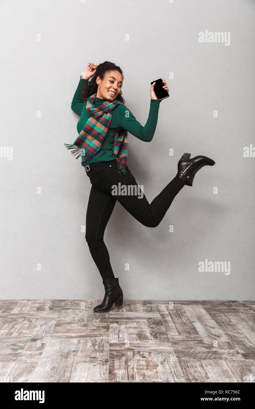 Full length portarit of a pretty african woman wearing scarf jumping over gray background, holding mobile phone Stock Photo