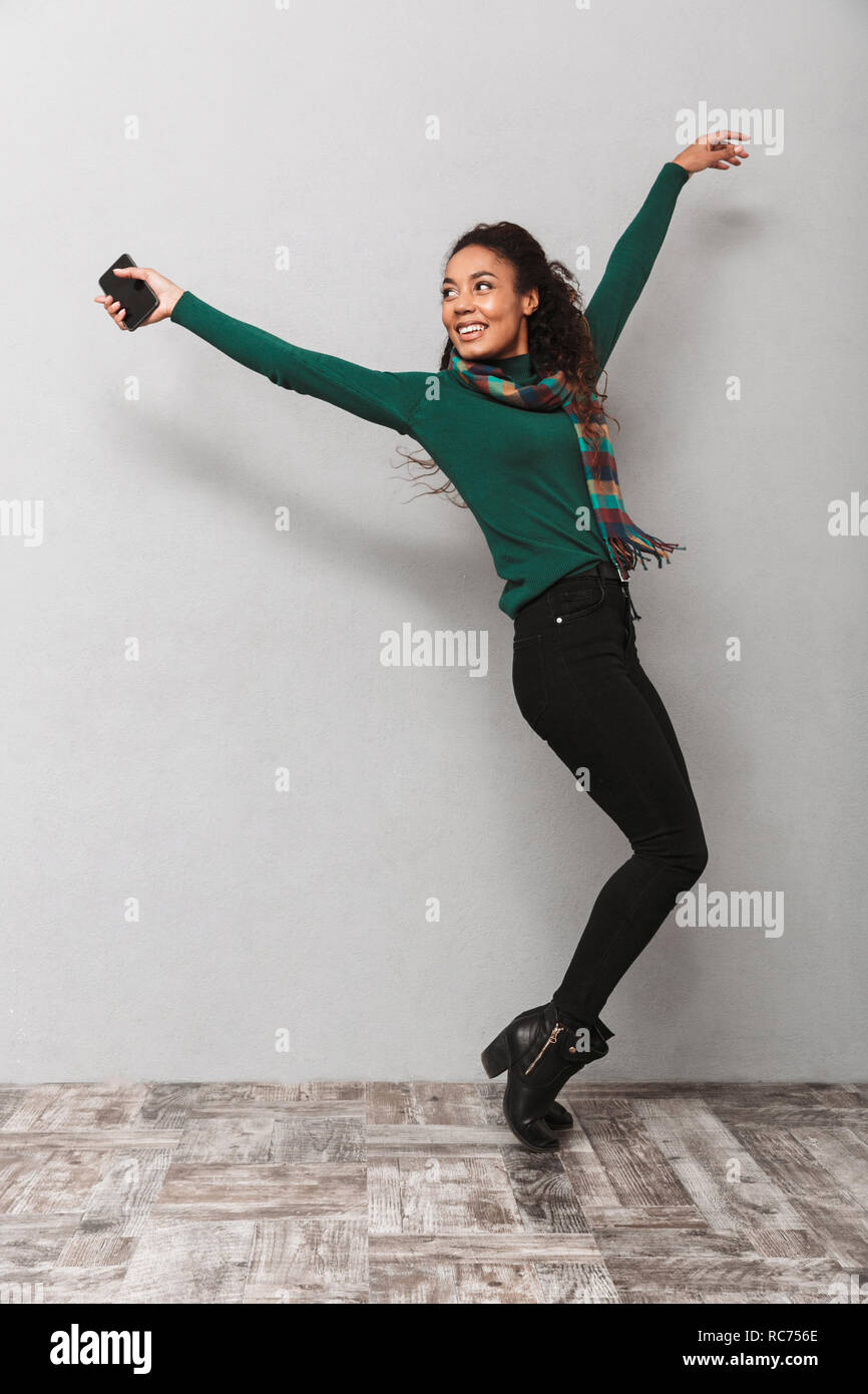 Full length portarit of a pretty african woman wearing scarf jumping over gray background, holding mobile phone Stock Photo