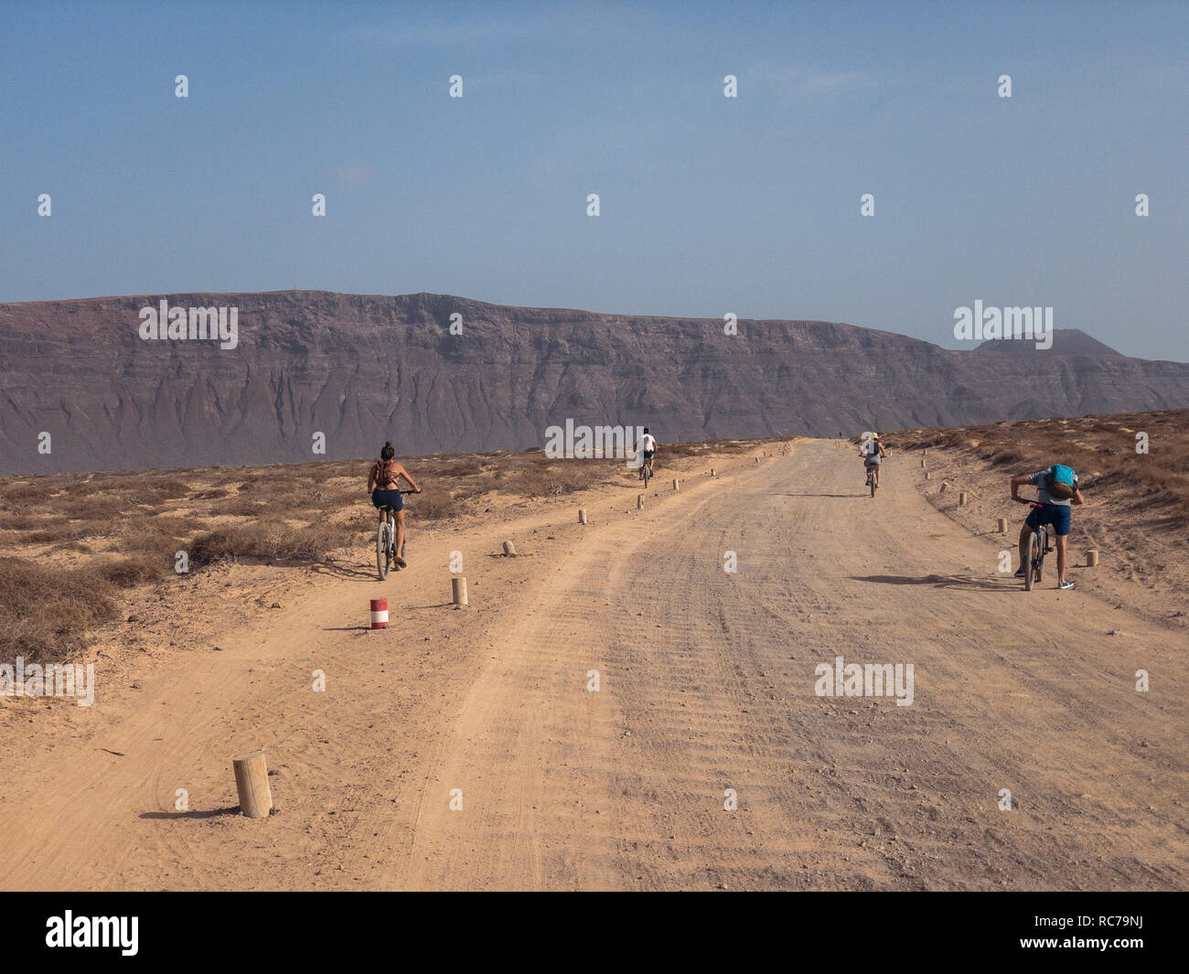 People cycling on a dirt path in the island of La Graciosa, Lanzarote, Canary Islands. Spain. In the background El risco, Lanzarote's mountain Stock Photo
