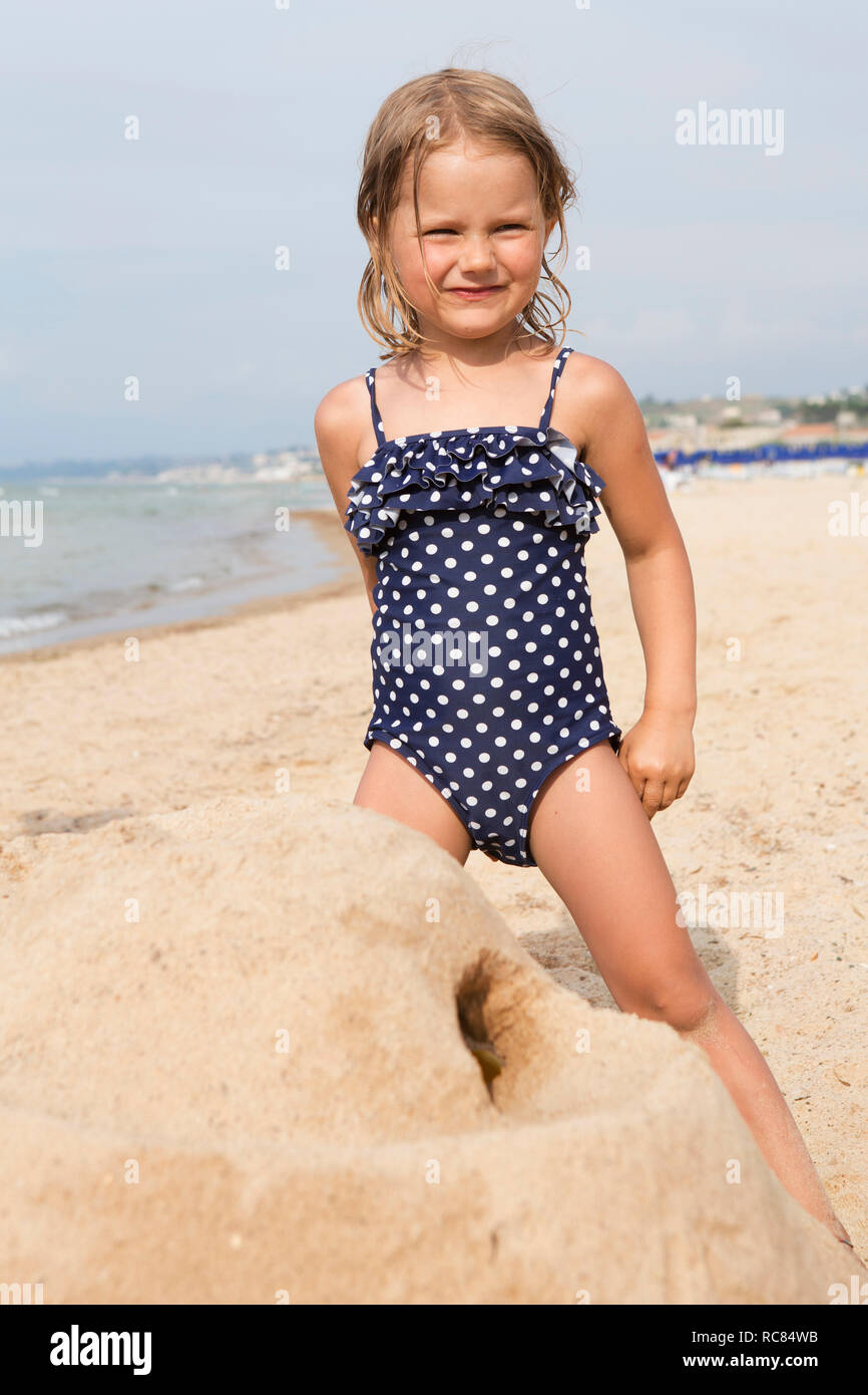 Cute girl building sandcastle on beach in spotted swimming costume, portrait, Castellammare del Golfo, Sicily, Italy Stock Photo