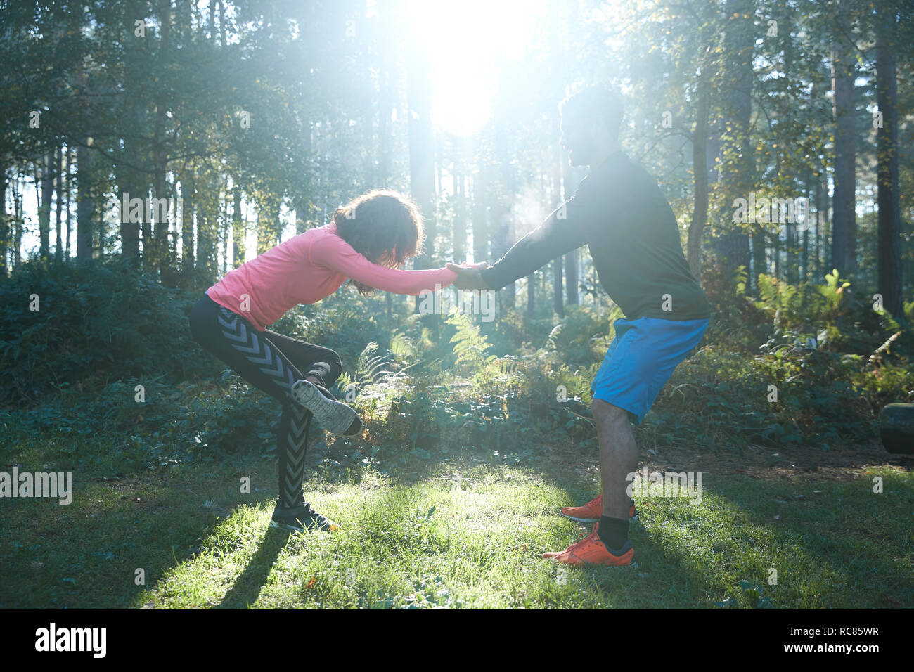 Male and female runners stretching legs and warming up in sunlit forest Stock Photo