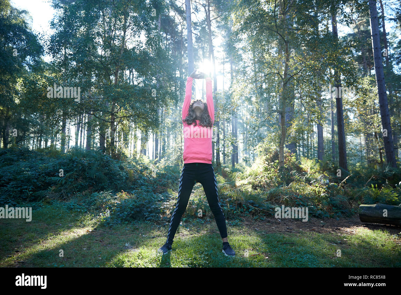 Female runner stretching arms and warming up in sunlit forest Stock Photo