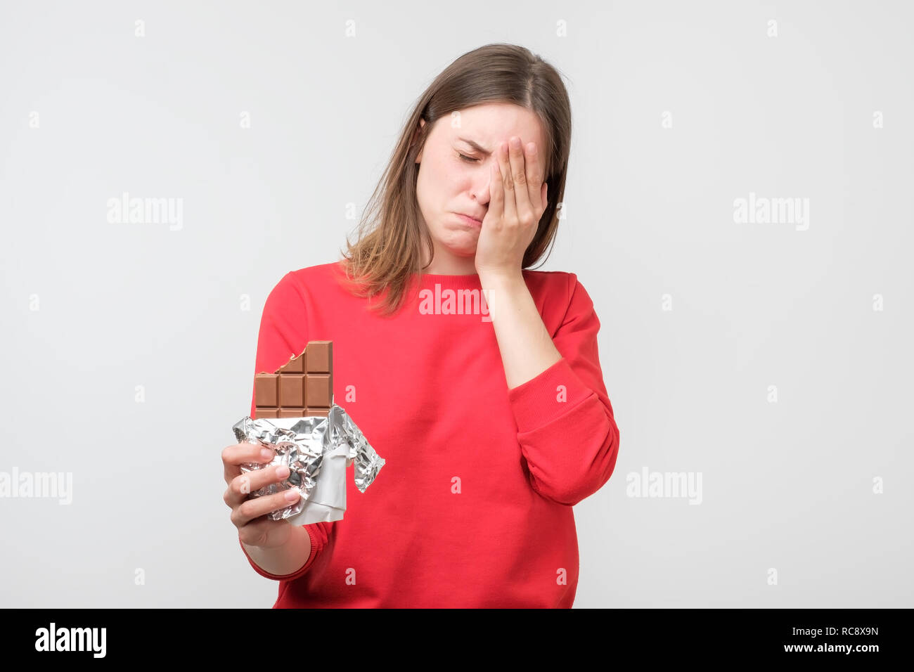 Sad young woman tired of diet restrictions holding sweet chocolate isolated on gray wall background. Nutrition concept. Feelings of guilt Stock Photo