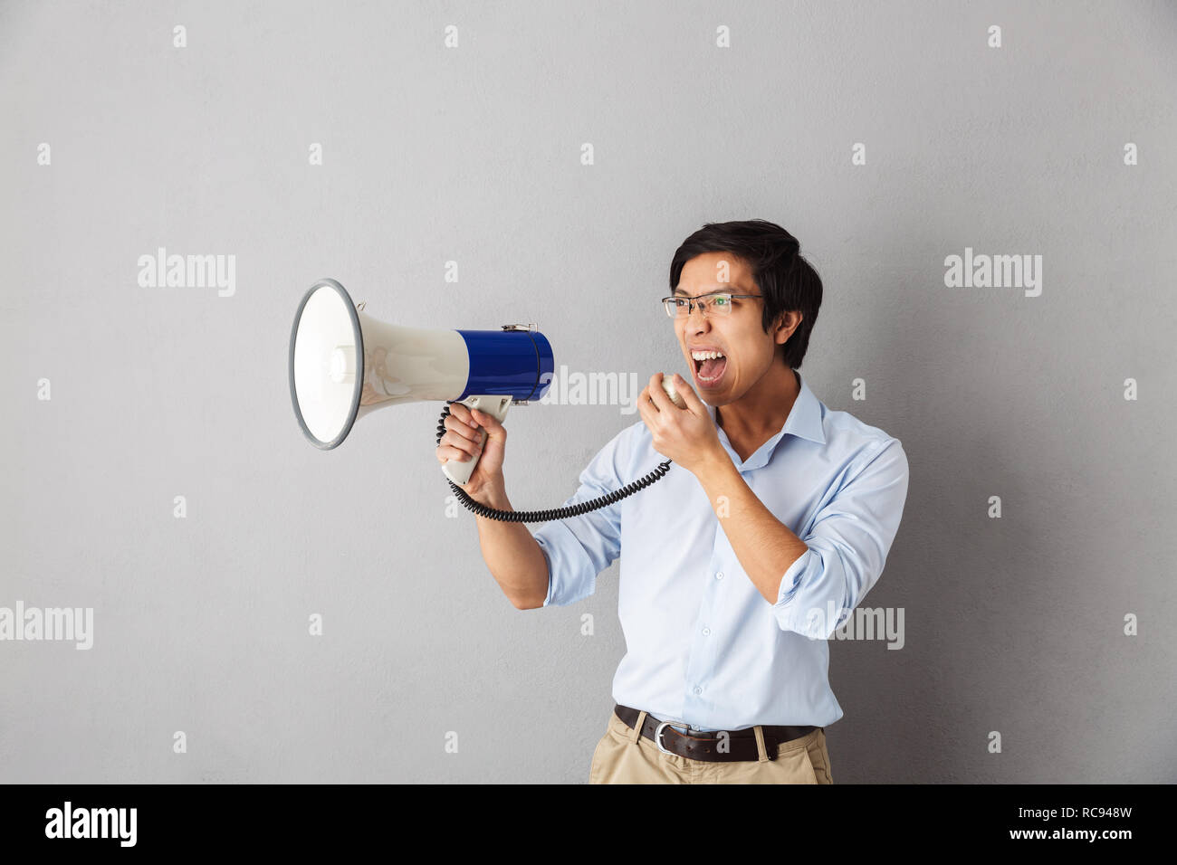 Angry asian business man standing isolated over gray background, screaming at a loudspeaker Stock Photo