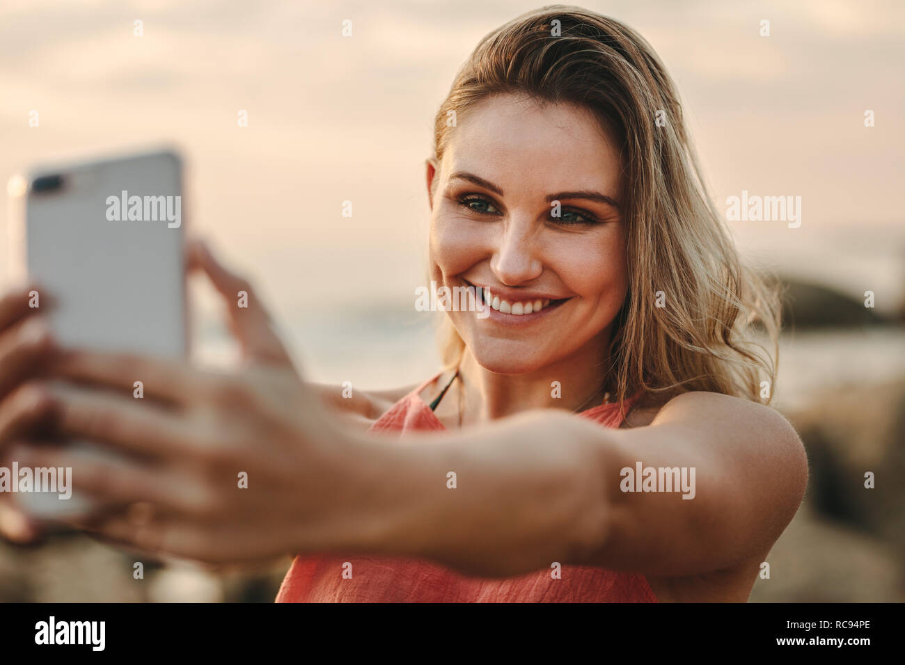 Smiling woman on vacation taking a selfie using mobile phone. Close up of a happy woman holding a cell phone and taking a selfie standing at the beach Stock Photo