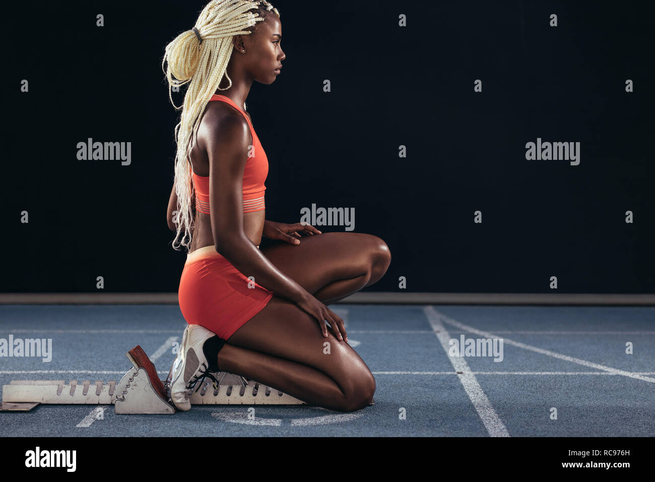 Female sprinter sitting at the start line on the starting block on a running track. Side view of female runner sitting at the start line on running tr Stock Photo