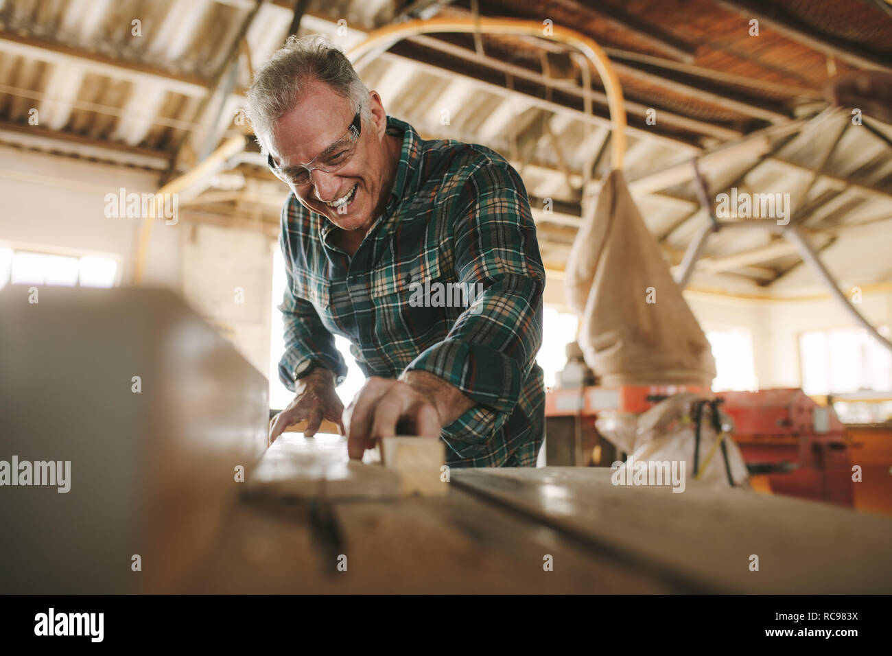 Happy senior male carpenter cutting wood planks on table saw machine. Smiling mature man working in carpentry workshop. Stock Photo
