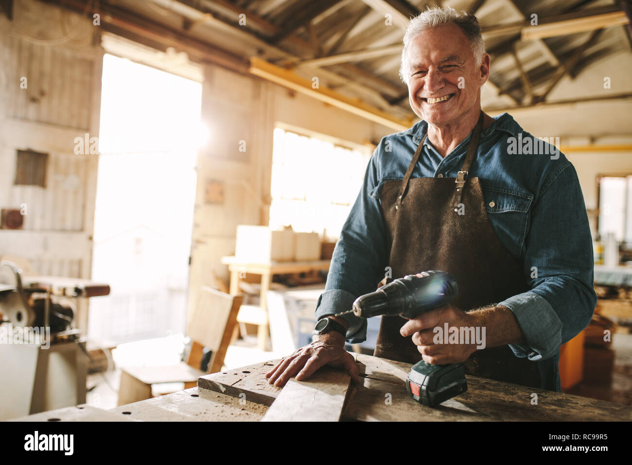 Smiling senior carpenter standing at workbench with a drill machine. Happy mature male carpenter at his workshop. Stock Photo