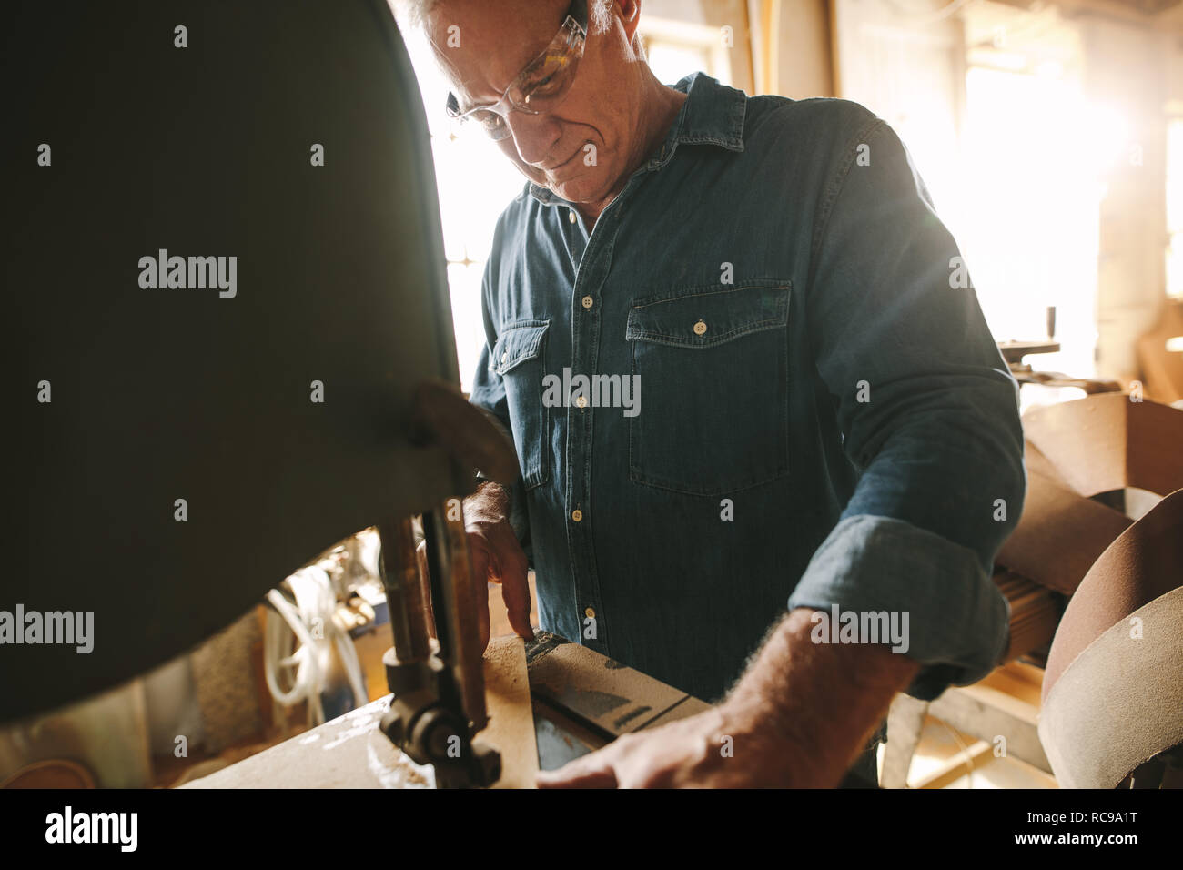 Senior male carpenter working on band saw machine cutting wood in his workshop. Carpenter cutting wood on machine at carpentry workshop. Stock Photo