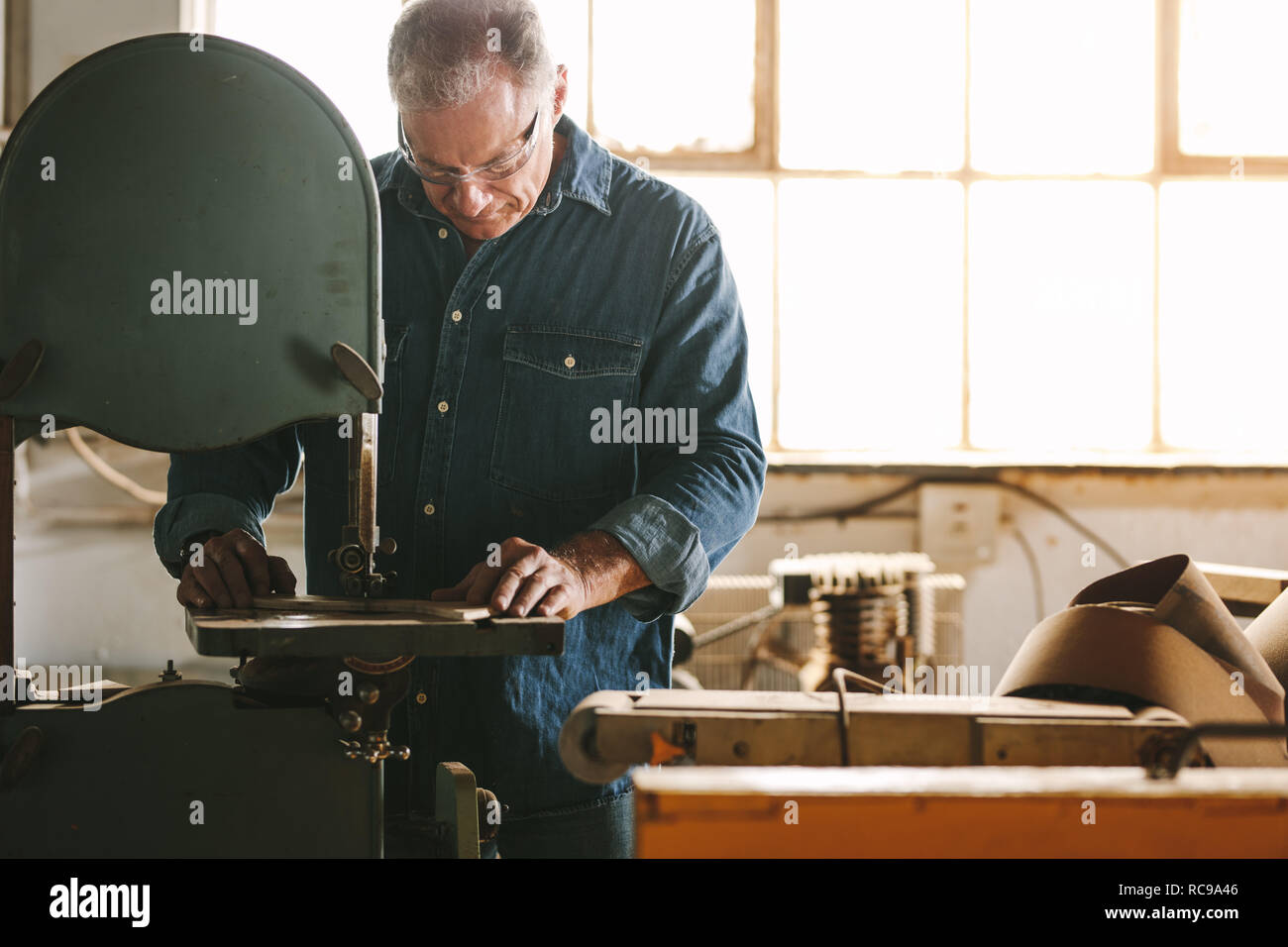 Mature worker in the carpentry workshop cuts the wood using band saw.  Carpenter working in carpentry preparing furniture parts. Stock Photo