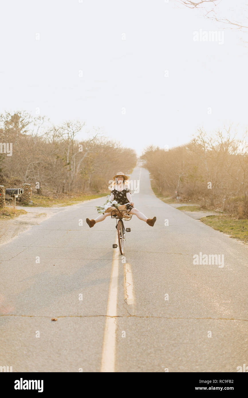 Young woman riding bicycle with legs raised on rural road, Menemsha, Martha's Vineyard, Massachusetts, USA Stock Photo