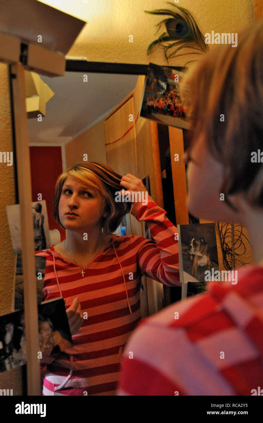 junge Frau zieht Grimassen vor dem Spiegel in ihrem Zimmer | young woman making funny grimaces in front of a mirror in her room Stock Photo