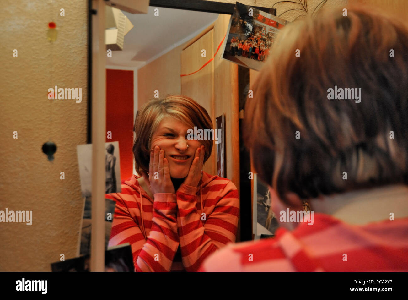 junge Frau zieht Grimassen vor dem Spiegel in ihrem Zimmer | young woman making funny grimaces in front of a mirror in her room Stock Photo