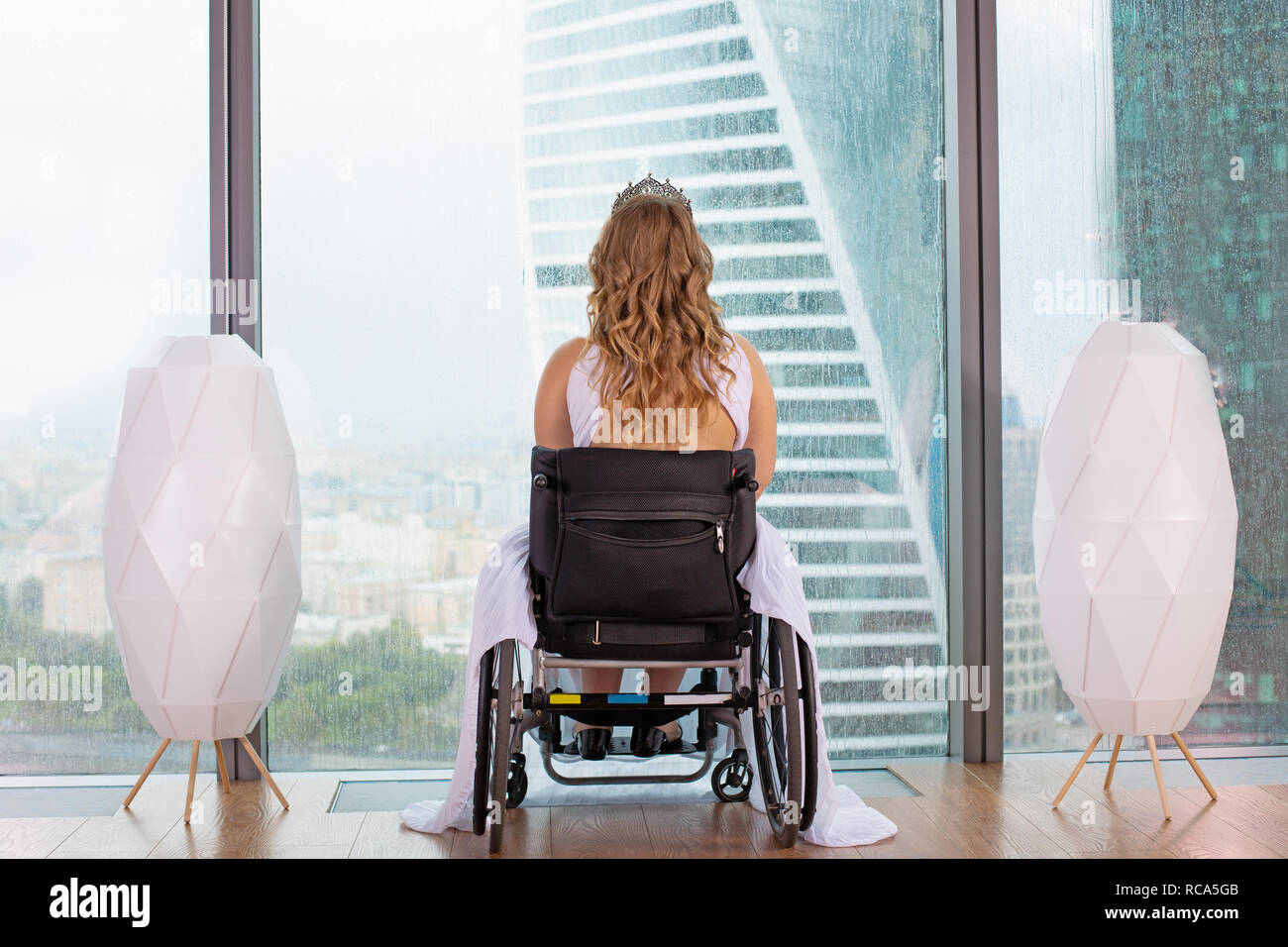 Beautiful disabled bride posing in a wheelchair against the background of a panoramic window overlooking the skyscrapers and a big city, back veiw, disability overcoming concept Stock Photo