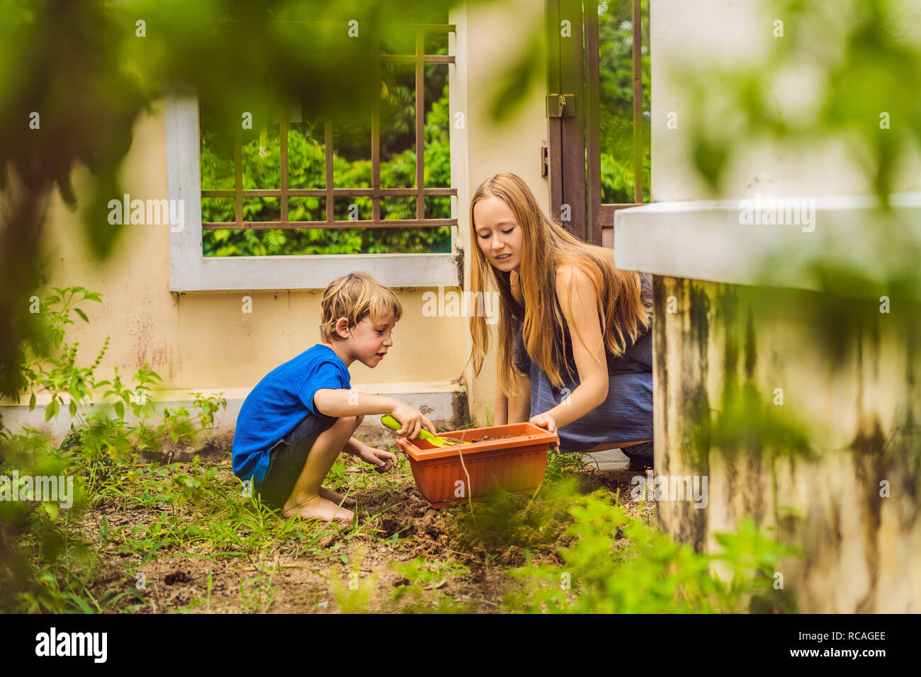 Beautiful young woman and her cute son planting seedlings in bed in the domestic garden at summer day. Garden tools, gloves and watering can outdoors. Gardening activity with little kid and family Stock Photo