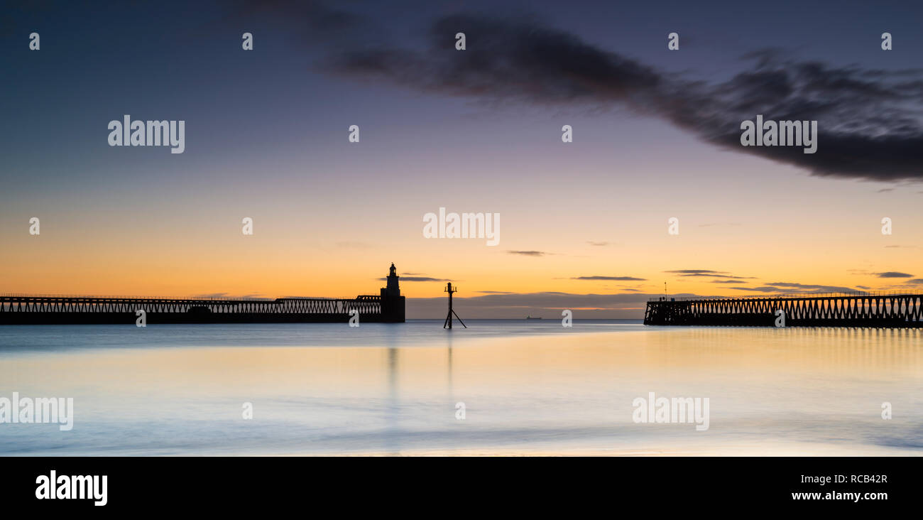 Winter sunrise at Blyth harbour looking across the mirror reflection on the tide towards the two silhouette piers and the lighthouse of the horizon Stock Photo