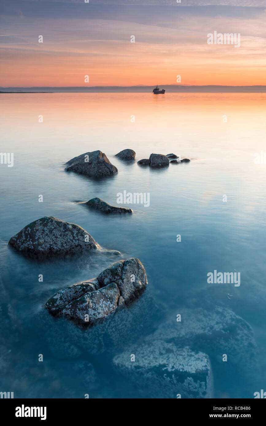 Calm peaceful sunset on Holy Island, lovely warm pastel colours in the sky & cool blues in the sea, the rocks standing proudly above & below the sea Stock Photo