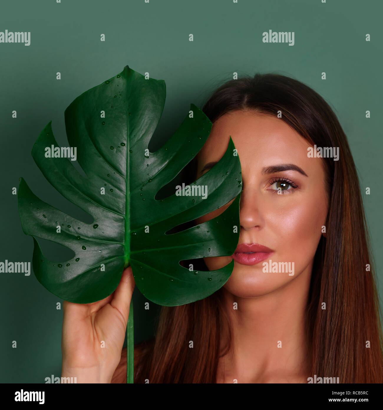 Young woman with natural make up and tropical monstera leaf in hand over green background with copy space. Skin care, pure beauty, cosmetics concept.  Stock Photo