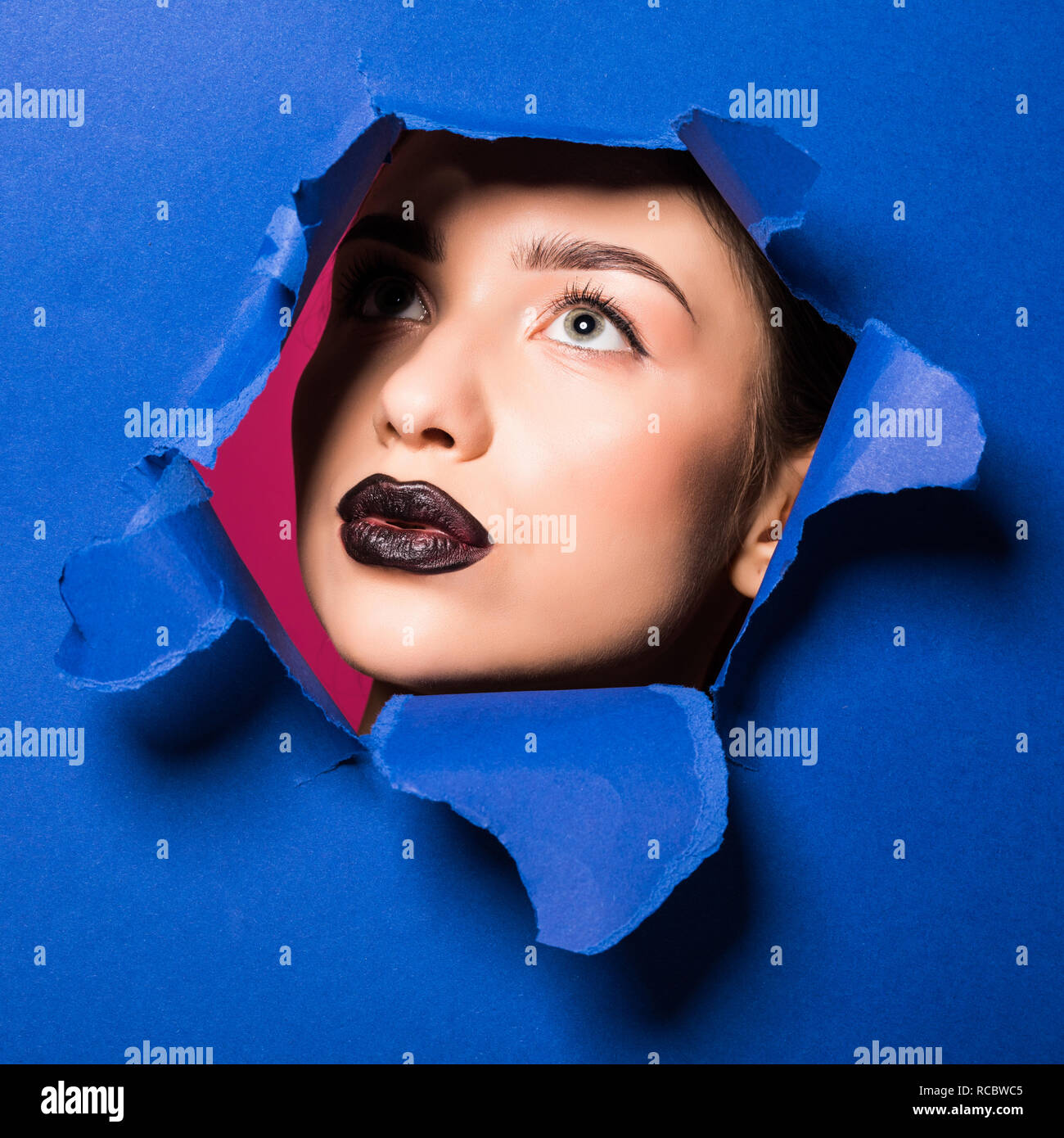 The Face Of Young Beautiful Girl With A Bright Make Up And Puffy Dark Lips Peers Into A Hole In Blue Paper Stock Photo Alamy