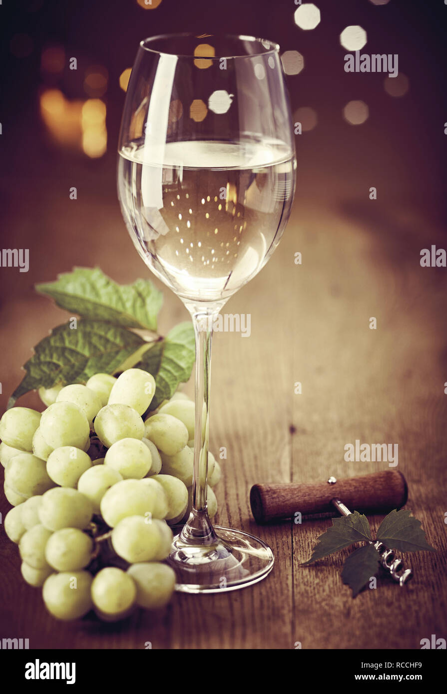 Elegant wineglass of white wine with bunch of fresh green grapes and bottle opener on a wooden table with a bokeh of sparkling party lights Stock Photo