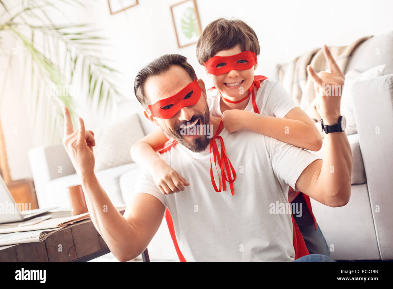 Father and little son wearing superheroe costumes together at home posing to camera man showing horns laughing cheerful Stock Photo