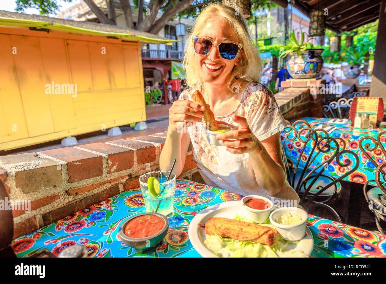 Joyful blonde woman puts enchilada in chili sauce, at typical Mexican food, at traditional restaurant of El Pueblo, Olvera street, Los Angeles Downtown State Historic Park, California, United States. Stock Photo