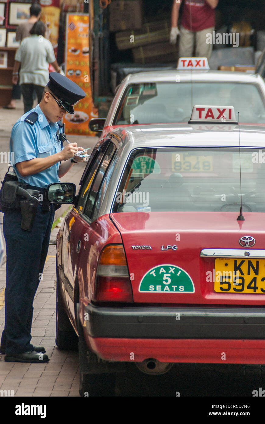 Hong Kong Island, China  - May 12, 2010: A traffic cop in blue writes a red taxi cab up for an infraction near Stanley Market. Faded shops and vendors Stock Photo