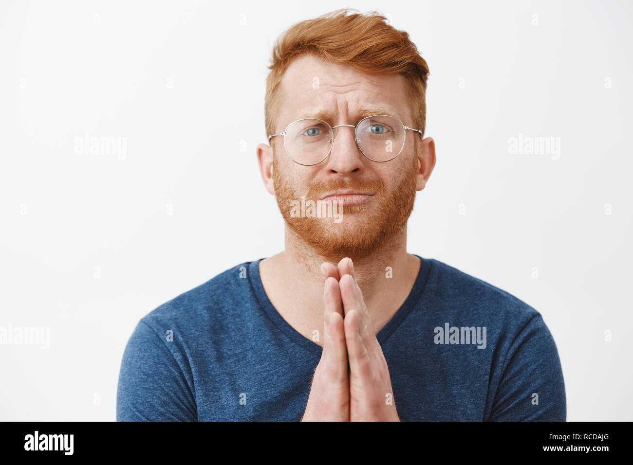 Close-up shot of redhead man in need standing with hands in pray over chest, pursing lips and frowning while asking forgiveness or begging for huge favour, being in troubled over gray background Stock Photo