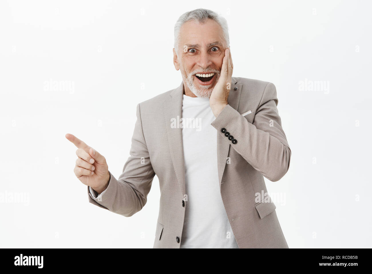 Waist-up shot of speechless delighted and surprised handsome elegant grandfather in stylish suit pressing hand to cheek from amazement and joy pointing left at curious thing smiling happily Stock Photo