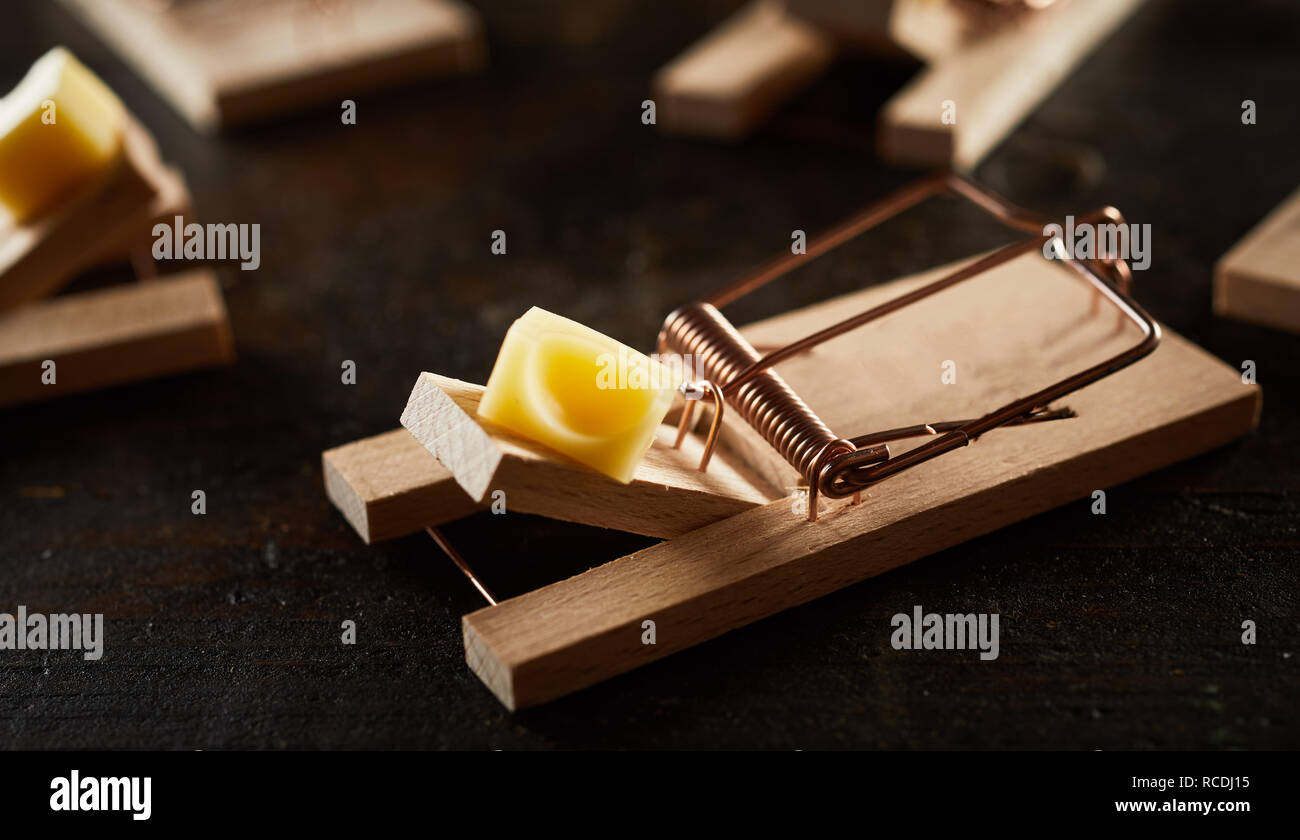 Cocked spring wooden mousetrap loaded with piece of cheese on wooden bar. Viewed in close-up among many, on dark surface Stock Photo