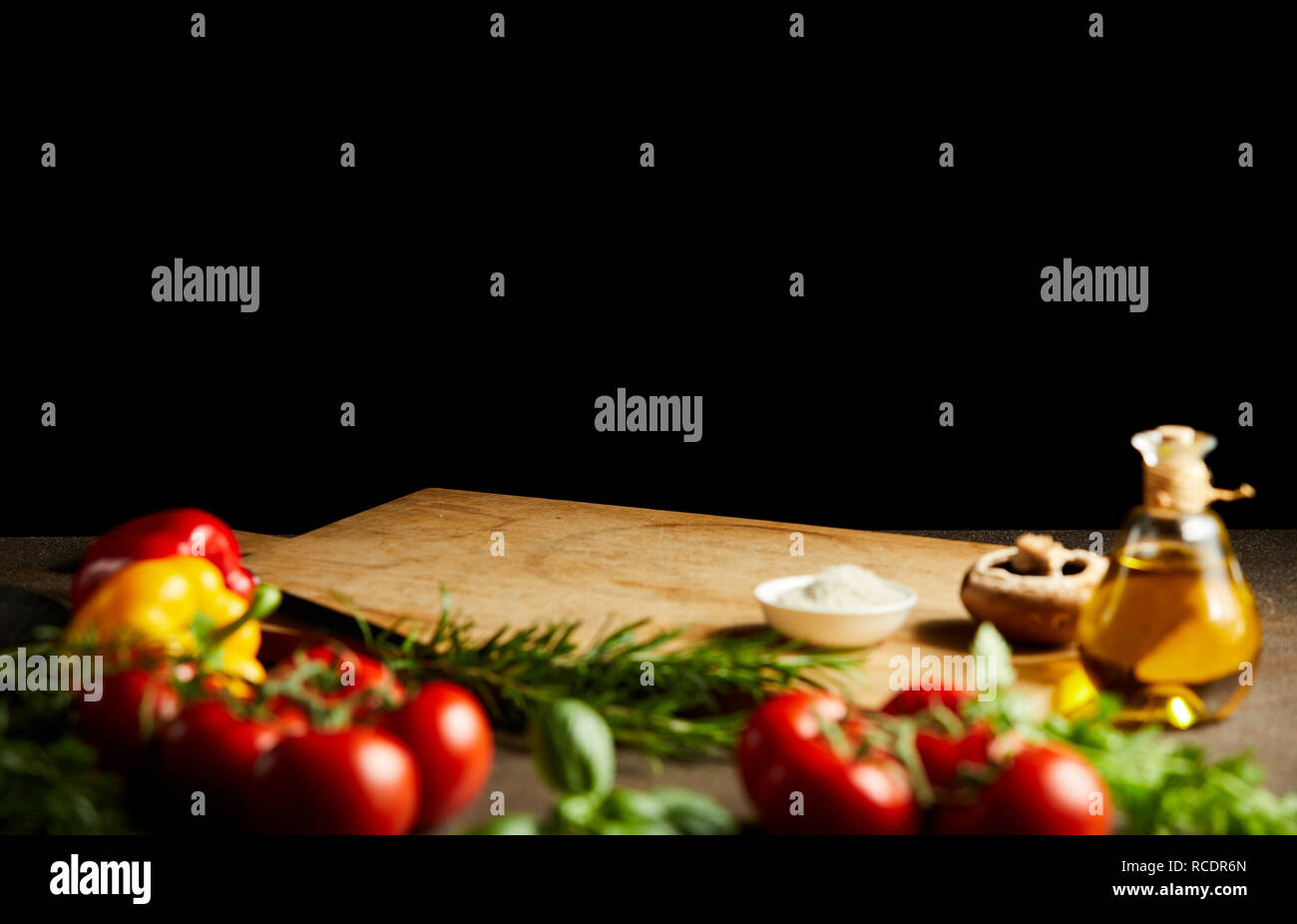 Fresh cooking ingredients around a wooden board with vegetables, herbs condiments and olive oil against a black background with copy space Stock Photo