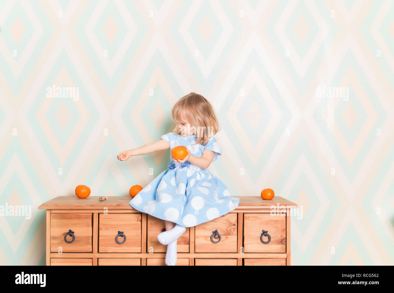 Portrait of blonde Caucasian baby girl with blue eyes in elegant dress and white socks sitting on wooden dresser with tangerine clementine fruit in hand and looking sideways at her hand. Amazement Stock Photo