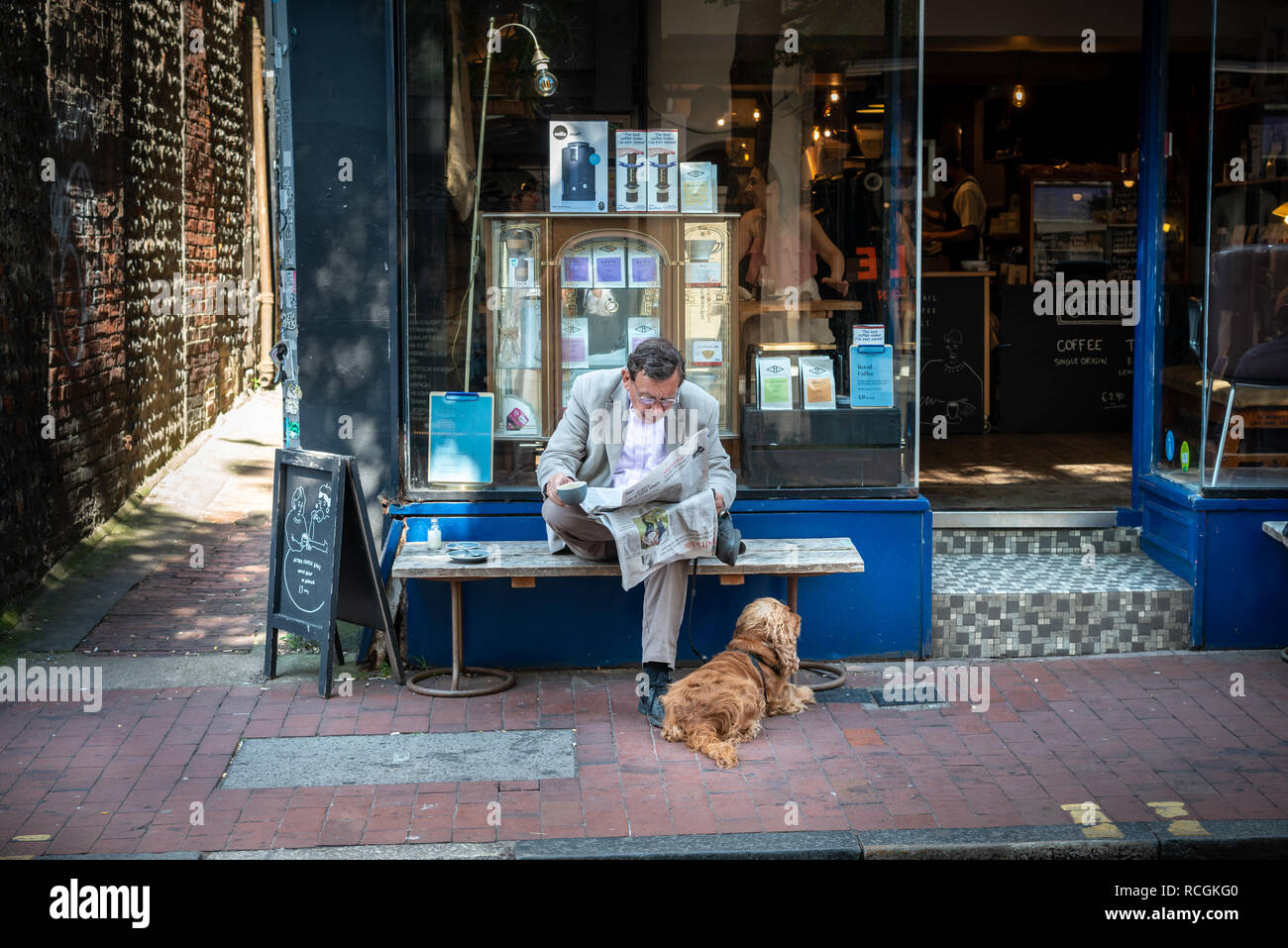 A man drinks coffee and reads a newspaper with a dog at his feet while sat outside a coffee shop in Brighton, East Sussex, England. Stock Photo