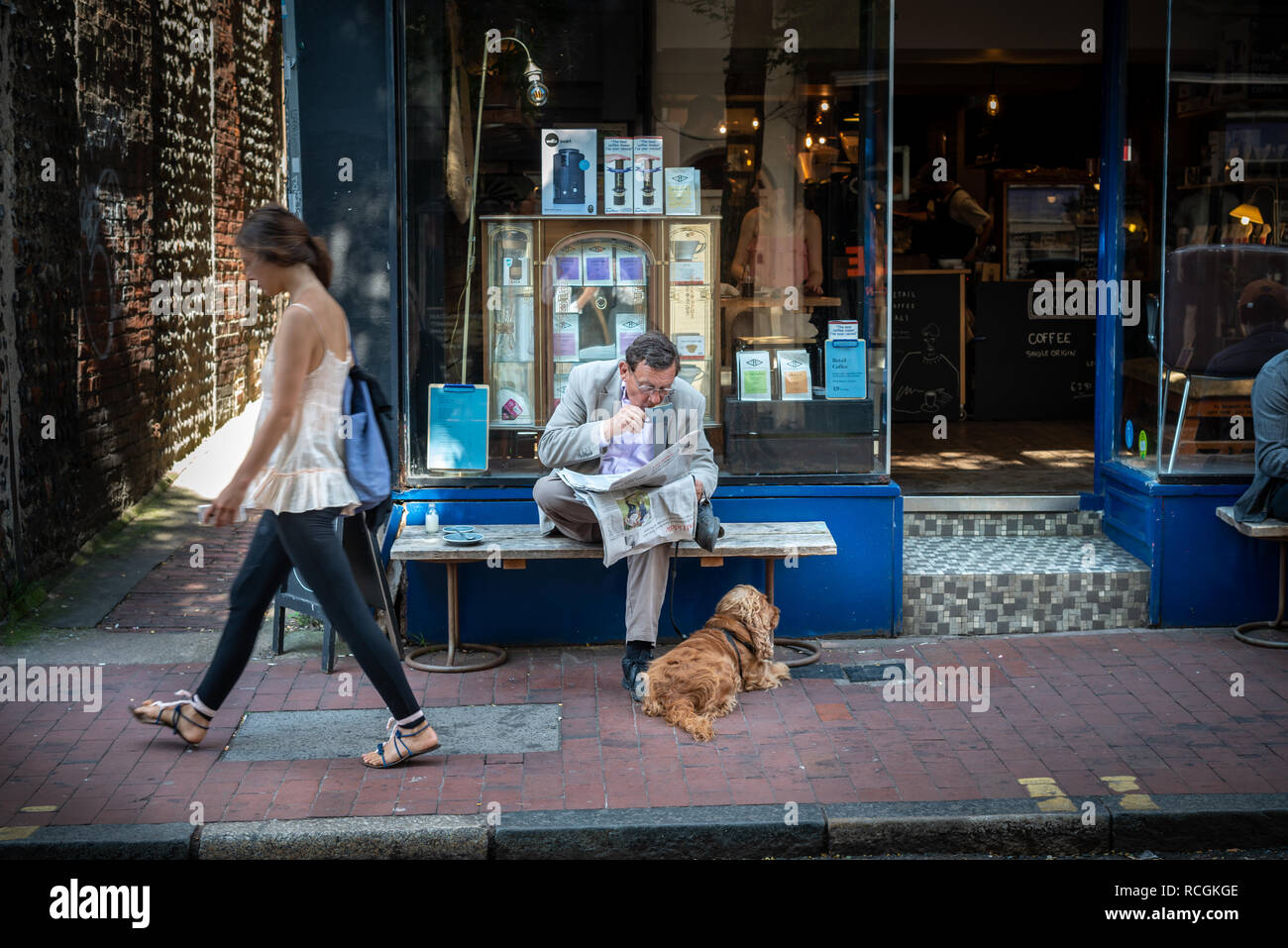 A man drinks coffee and reads a newspaper with a dog at his feet while sat outside a coffee shop in Brighton, East Sussex, England. Stock Photo