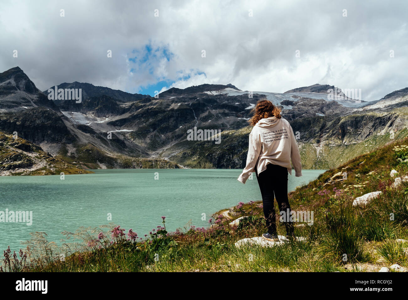 Young woman looking at Alpine lake with space for copy. She is curly and wears casual clothes. Rear view. Stock Photo