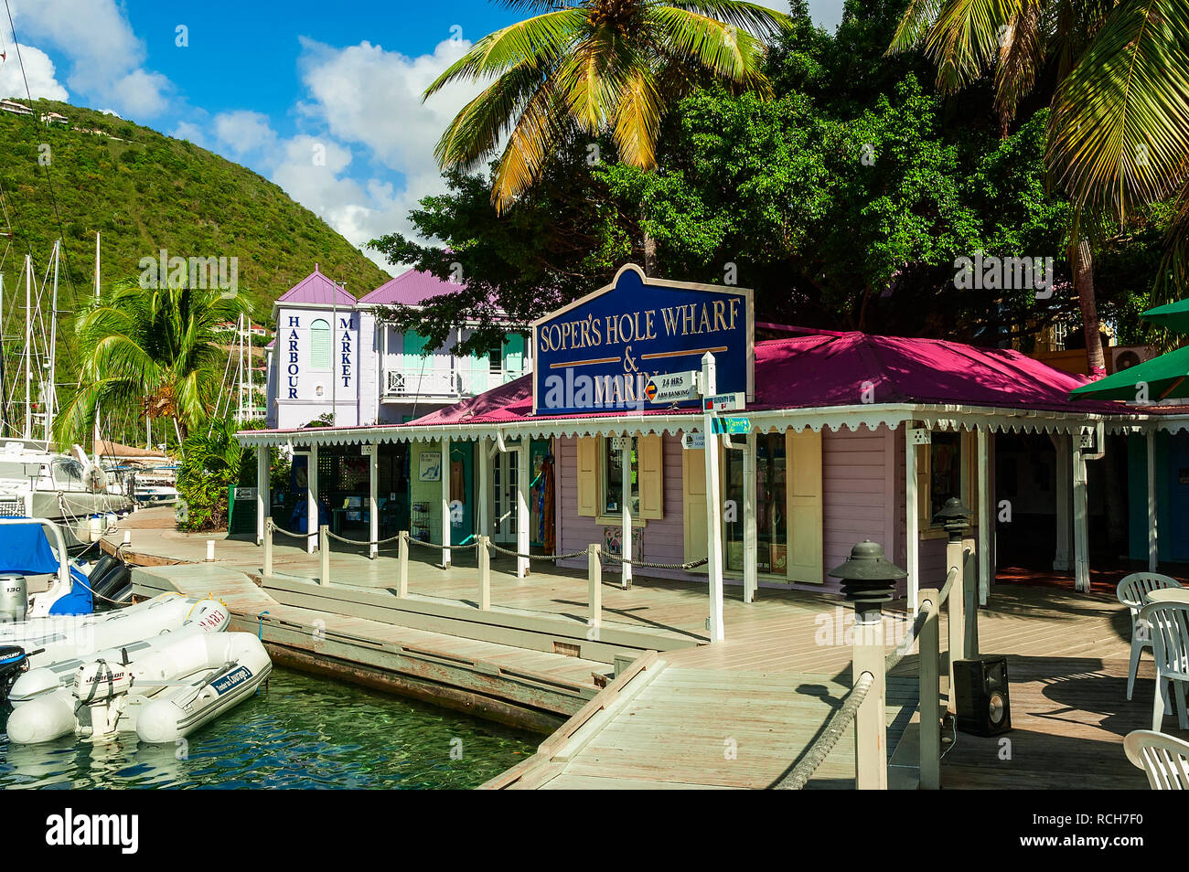 Sunshine bathes a bell shaped green, tree covered mountain, standing as a backdrop to the pristine buildings, boardwalk and dinghies at Soper's Hole Stock Photo