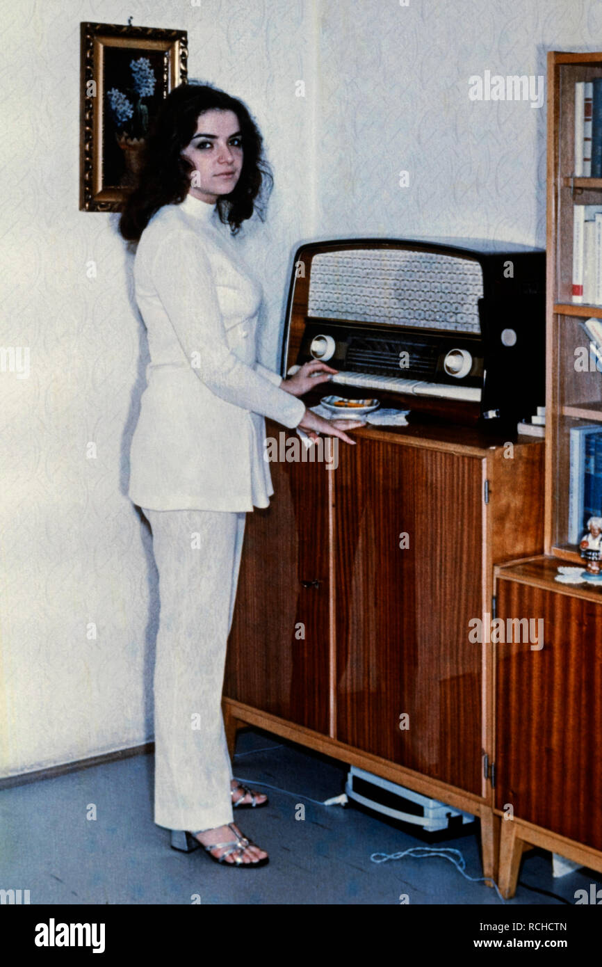 young pretty brunette female wearing a white trouser suit standing next to vintage radio set 1970s hungary Stock Photo