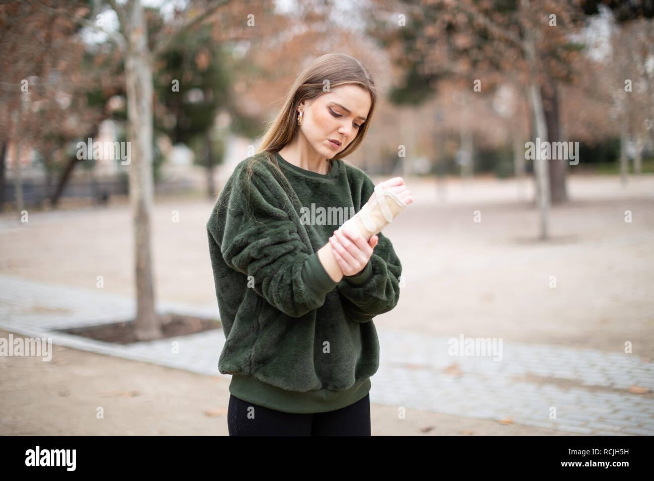 Beautiful young lady in stylish outfit frowning and touching bandage on injured arm while standing in autumn park Stock Photo