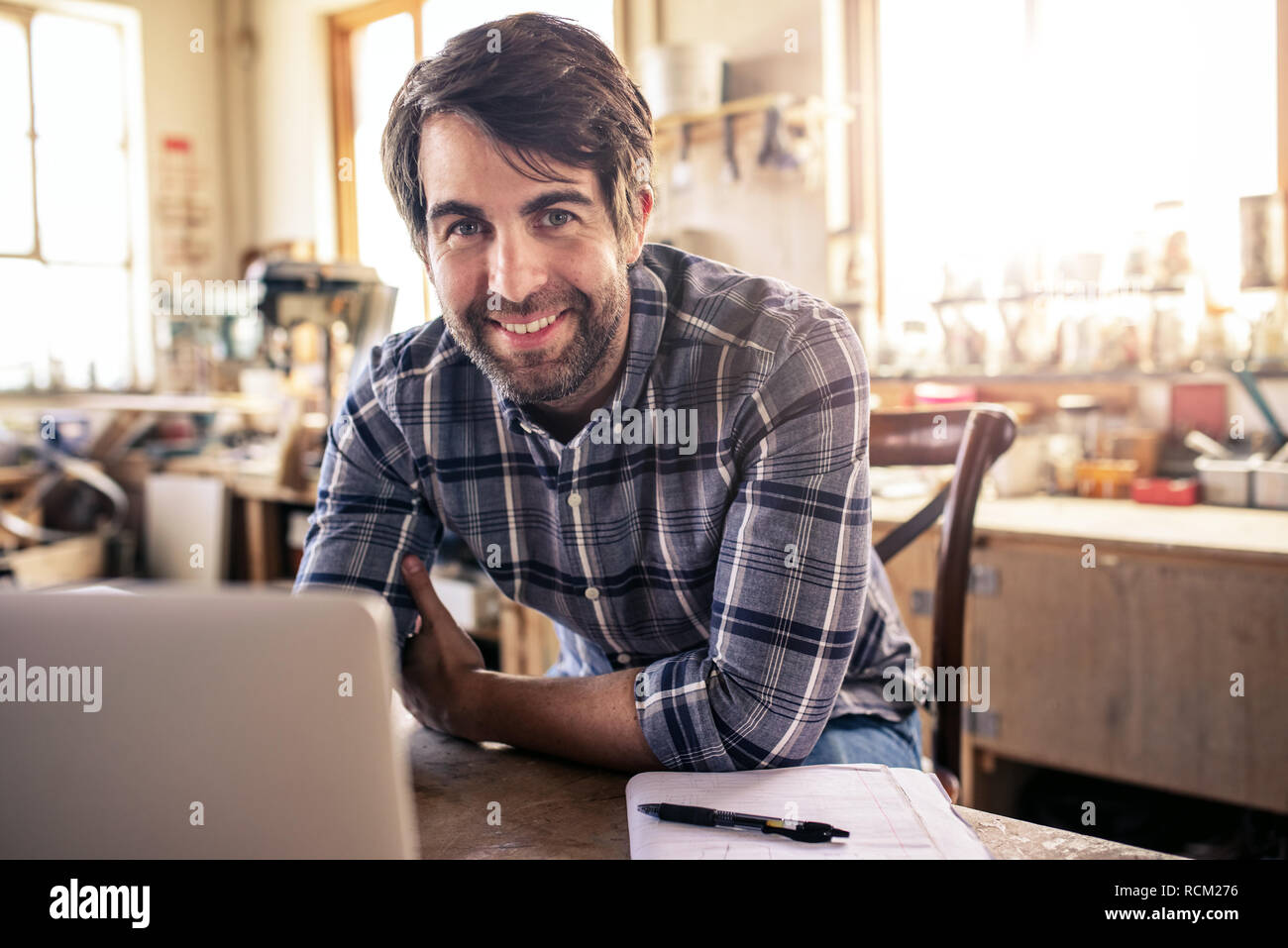 Woodworker smiling while using a laptop in his workshop Stock Photo