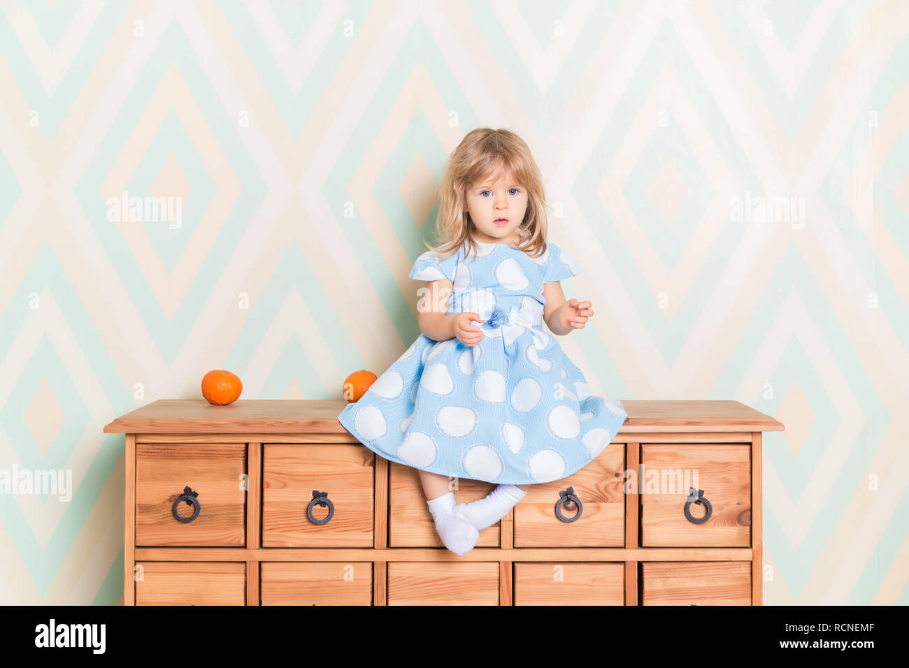 A little baby girl in her room sitting cross-legged on chest of drawers with tangerines on the rhomb wallpaper background. Child in blue polka dot dress and white socks attentively looking at camera. Stock Photo
