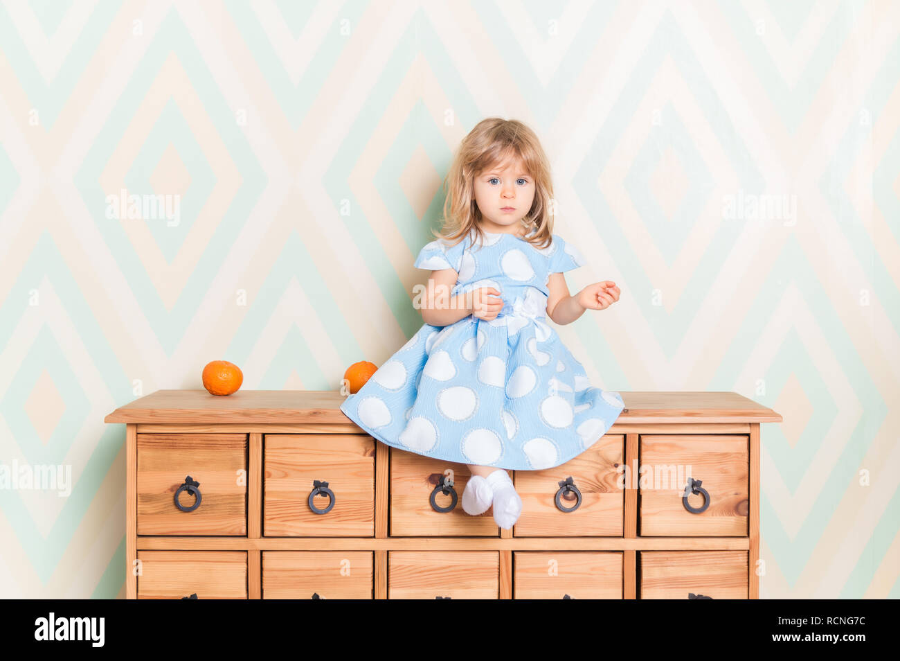 A little baby girl in her room sitting cross-legged on chest of drawers with tangerines on the rhomb wallpaper background. Child in blue polka dot dress and white socks attentively looking at camera Stock Photo