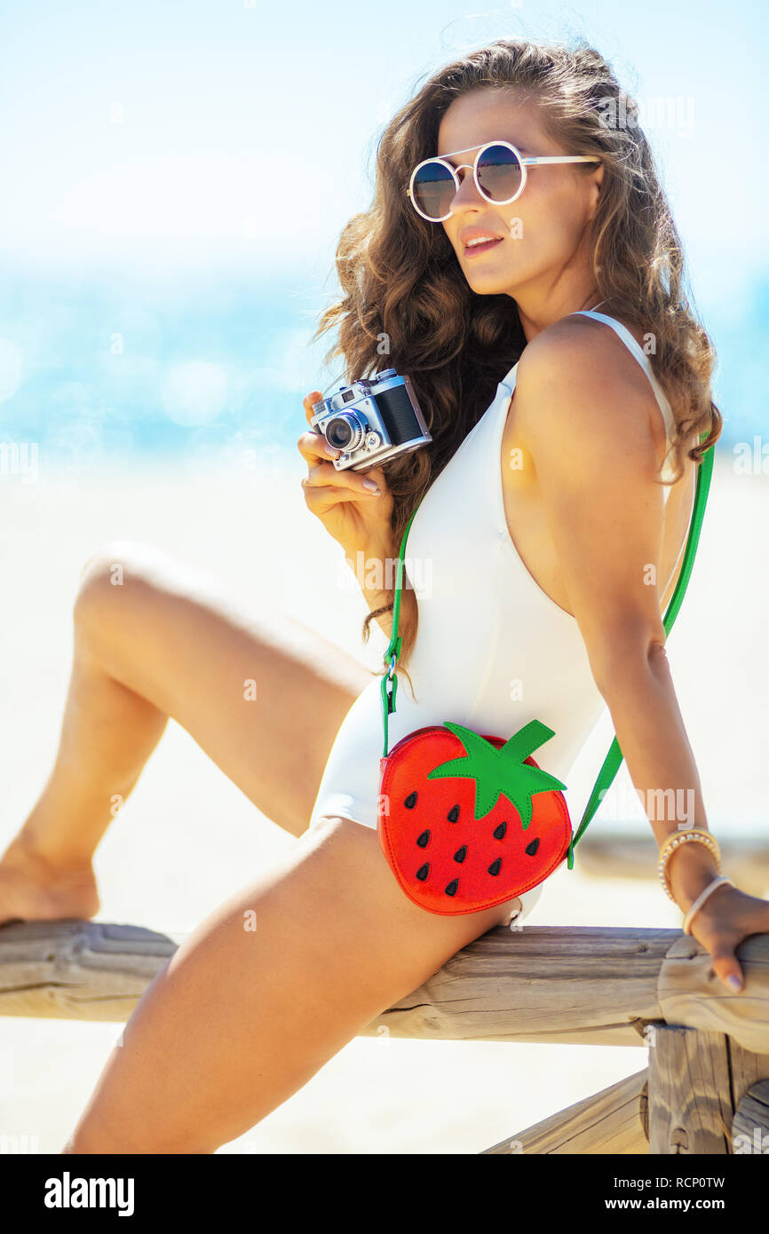 trendy woman in white beachwear with retro photo camera sitting on a wooden fence on the seacoast. woman in UVA UVB protectant sunglasses with funny s Stock Photo