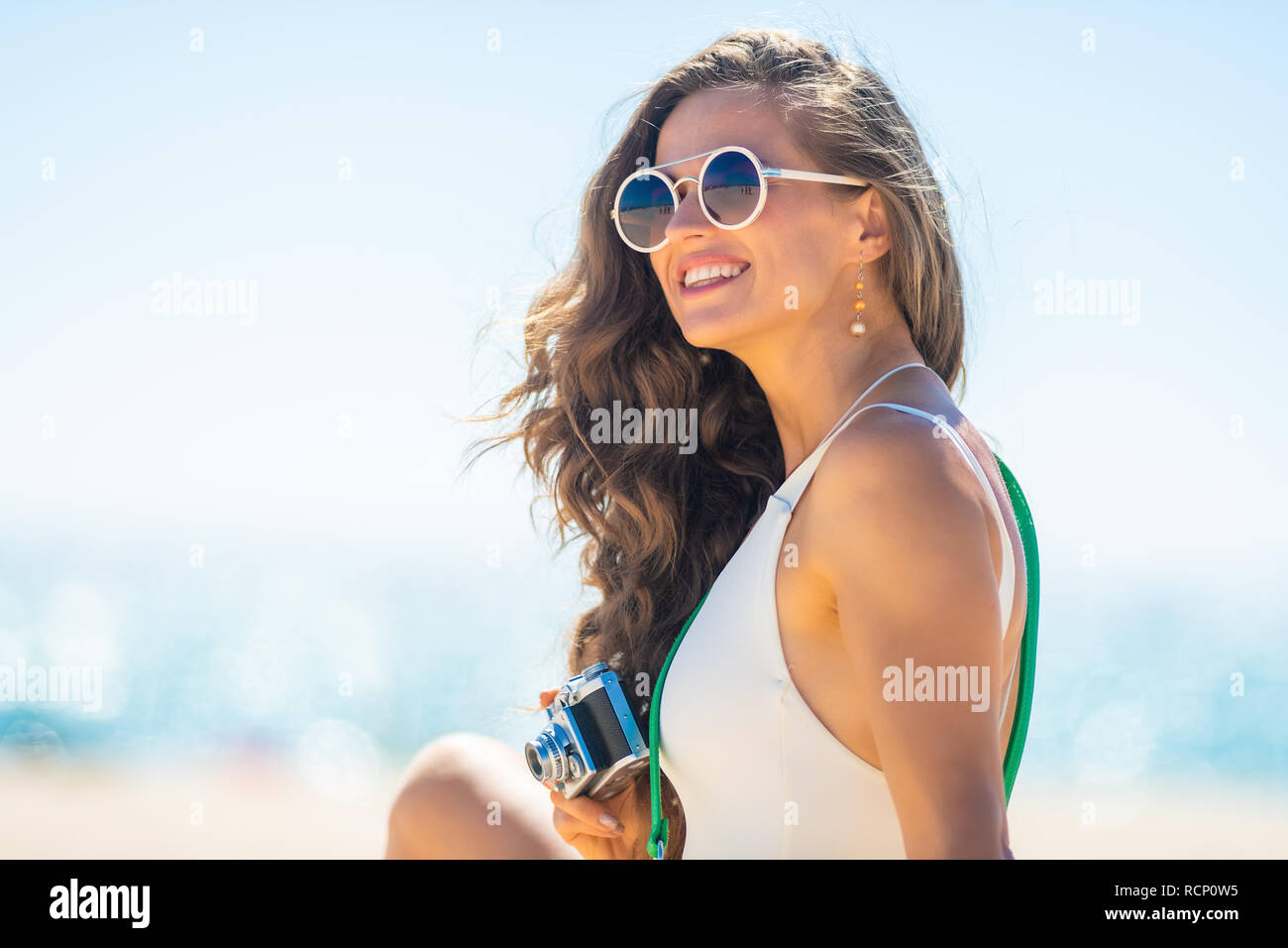Portrait of smiling trendy woman in white beachwear with retro photo camera looking into the distance on the beach. woman in UVA UVB protectant sungla Stock Photo