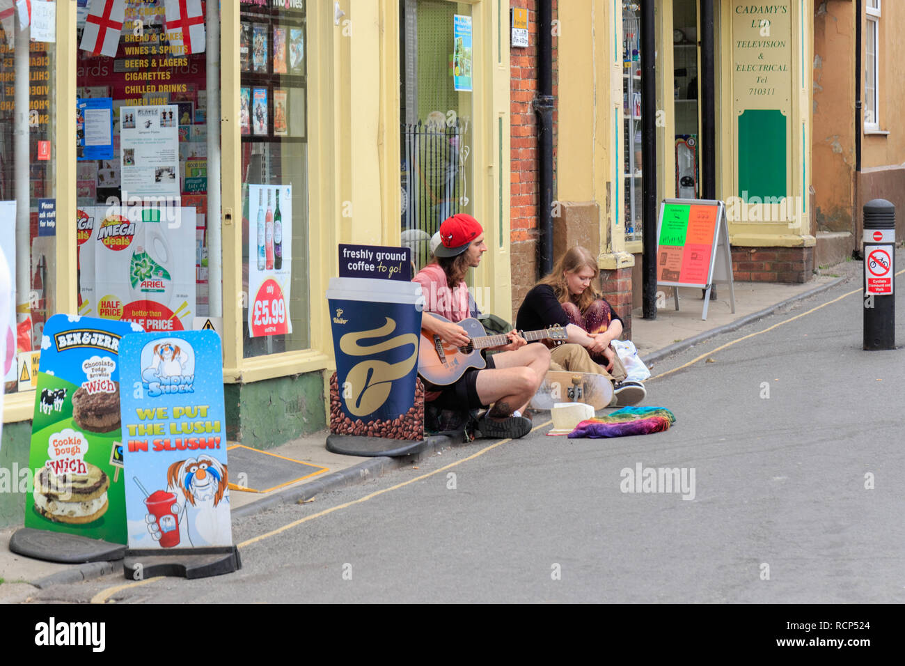 Male busker sitting on the pavement outside a shop playing his guitar. Summer 2018. Wells-next-the-sea, Norfolk, England UK Stock Photo